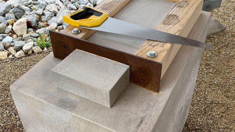 A handsaw lies on top of wood blocking that holds a stone slab in place on top of a large stone base. 