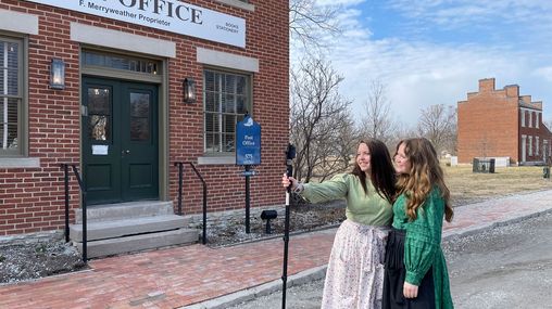 Two young women stand in the road in front of a brick building while smiling toward a mounted smart phone that one of them is holding up. 