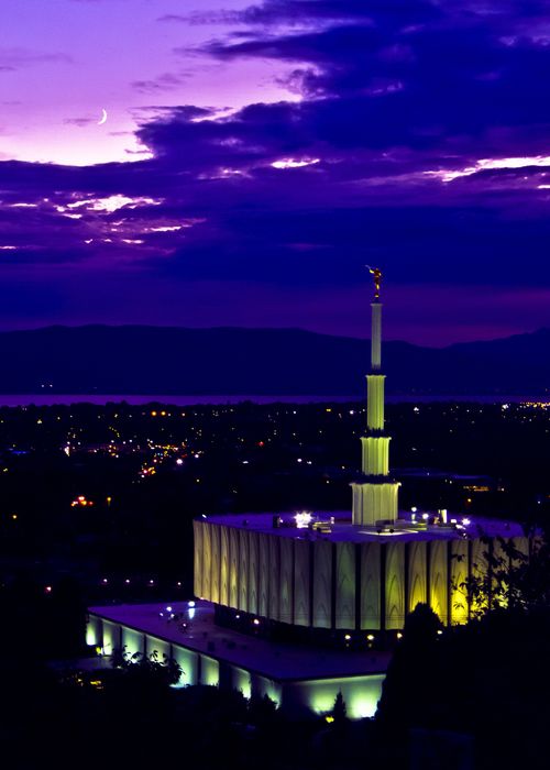 The Provo Utah Temple lit up at night, with a view of the city below, including a partial view of the lake in the background.
