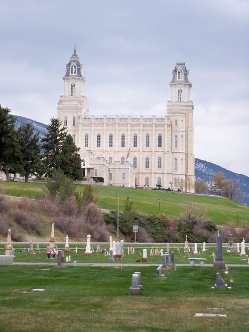 The Manti Utah Temple is on a hill, with the gravestones of a cemetery below, on an overcast day.