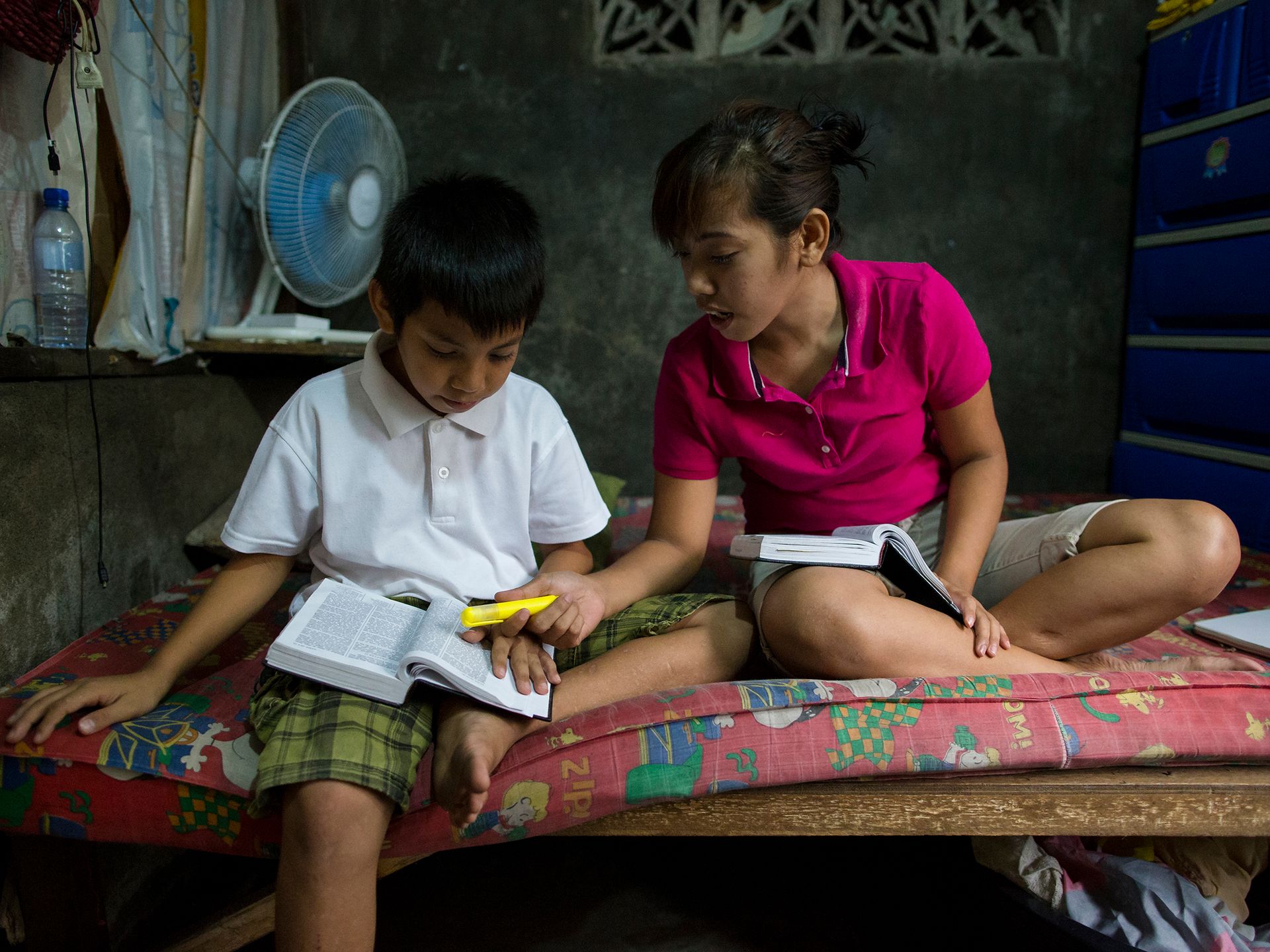 Family members in the Philippines studying together.  Mother and daughter study together and two children sit on a bed studying. They are studying scriptures and other books.