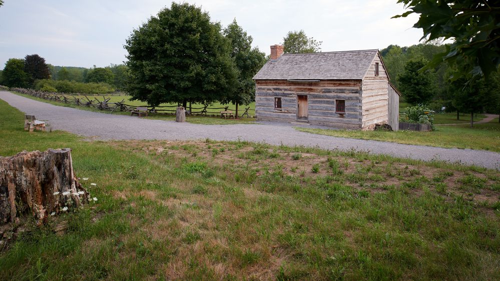 An exterior view of Joseph Smith’s family home in Palmyra, New York. The home is made of logs and has a field around it with trees behind.