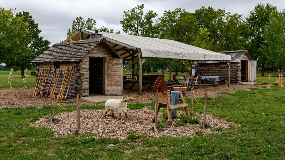 Covered pavilion with two small log cabins on both sides. A play horse and sheep are in the foreground.