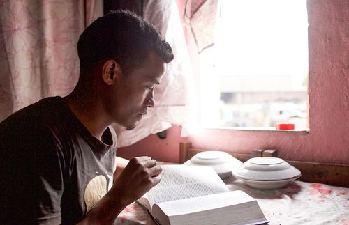 a young adult male reads the scriptures next to an open window