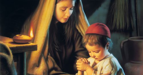 Jesus Christ depicted as a child with his mother Mary. Christ is kneeling beside Mary and resting His clasped hands on Mary’s lap. Mary has her head bowed as she and the young Christ pray. There is an oil lamp burning on a table next to the mother and son.