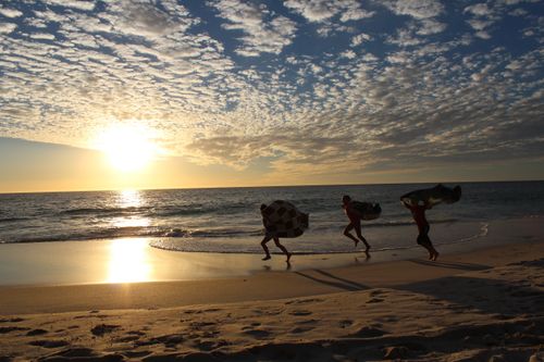 Silhouettes of three children carrying towels in the wind and running along the beach, with the sun starting to set behind them.