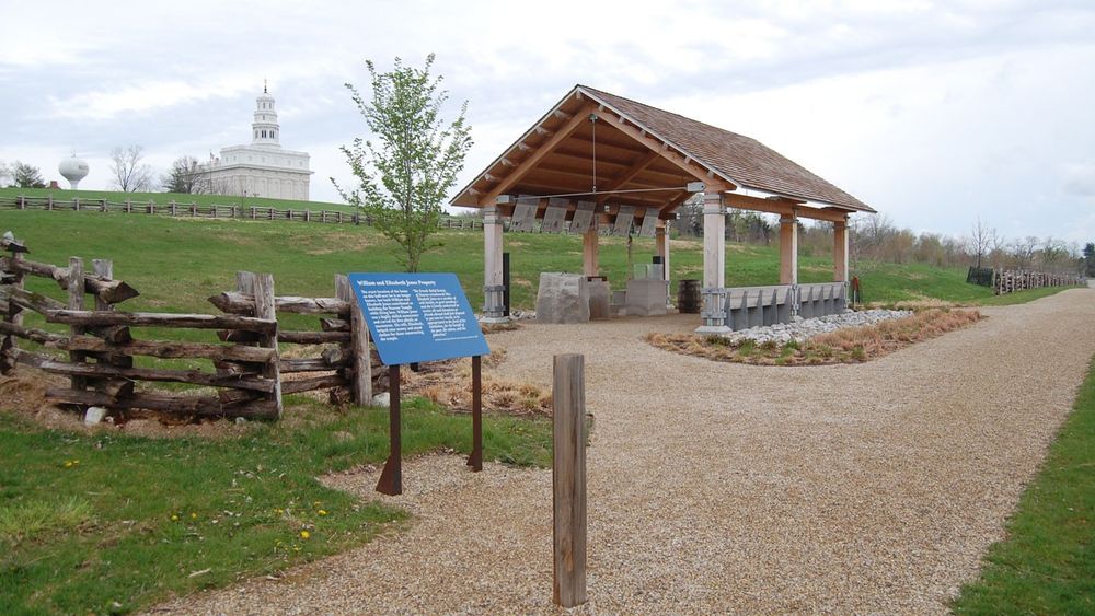 A modern, open-air pavilion made of wood with large stone blocks and signs mounted overhead underneath the roof with a white stone temple uphill in the background.