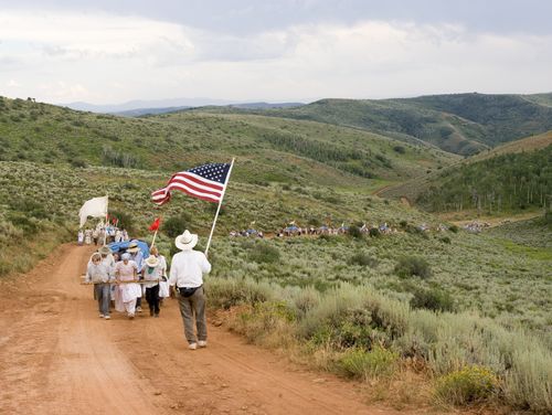 A man stands and waves an American flag as he walks in front of a large group of men and women dressed as pioneers pulling handcarts up a green hill.