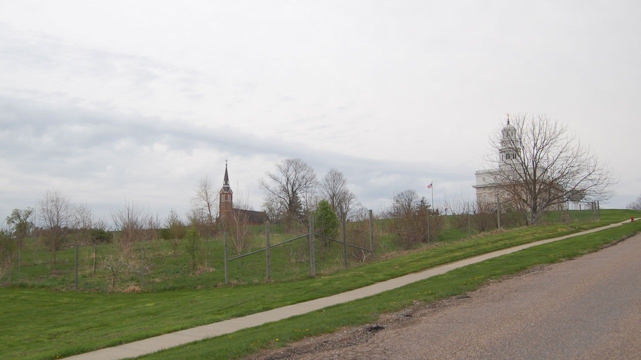 A fenced-in grouping of small trees with a brick church and white stone temple uphill in the background. 