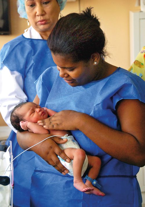 Two female doctors in blue vests, one checking the breathing of a newborn baby, in a neonatal intensive care unit in the Dominican Republic.