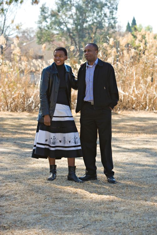 A man in a plaid shirt, jacket, and dress pants standing next to his wife in a jacket, skirt, and boots outside in Africa.