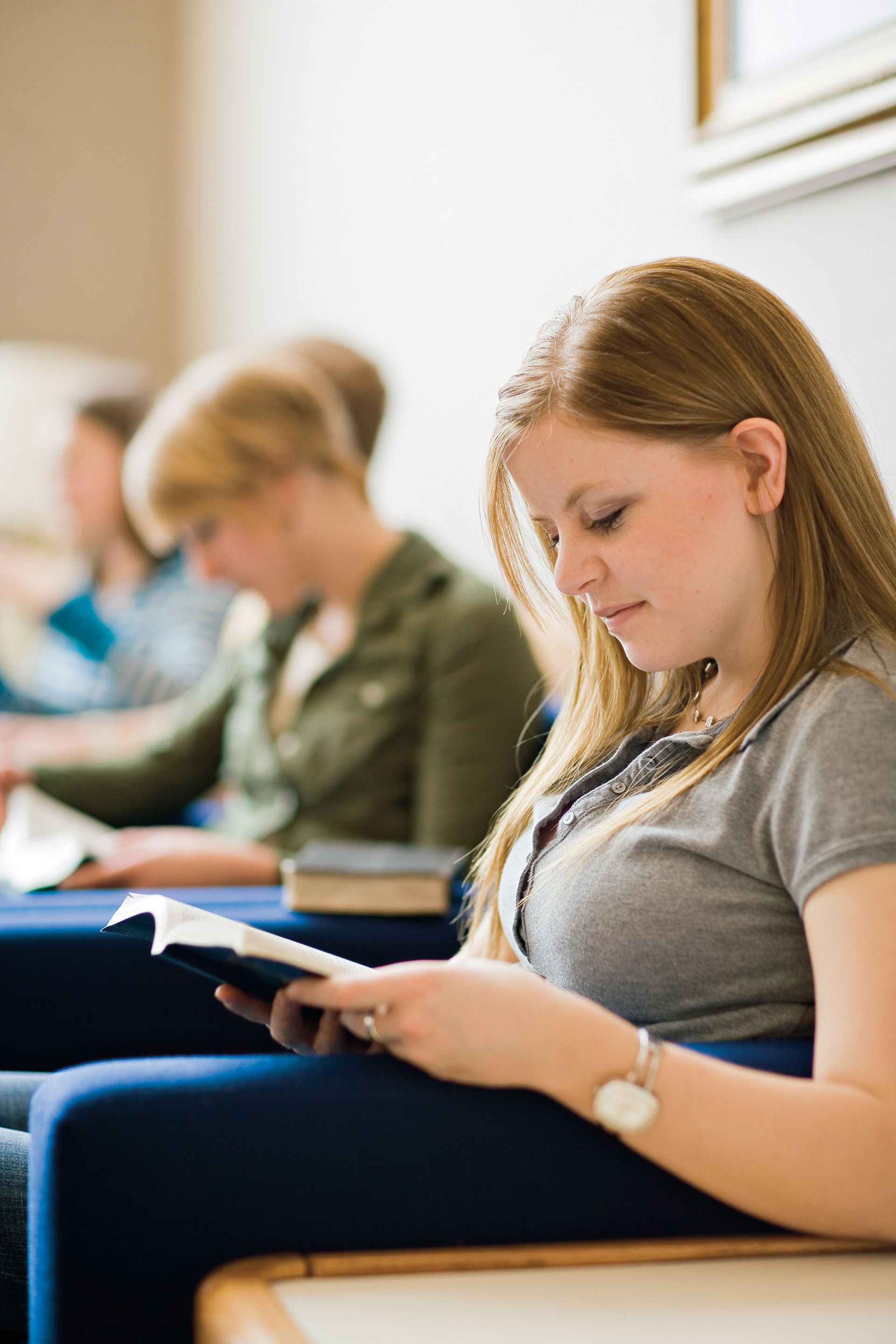 A young woman sits in a chair and reads from a set of scriptures, with other young adults around her.