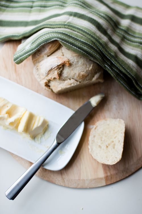 A fresh loaf of bread under a tea towel, on a cutting board next to a plate of butter and a knife.