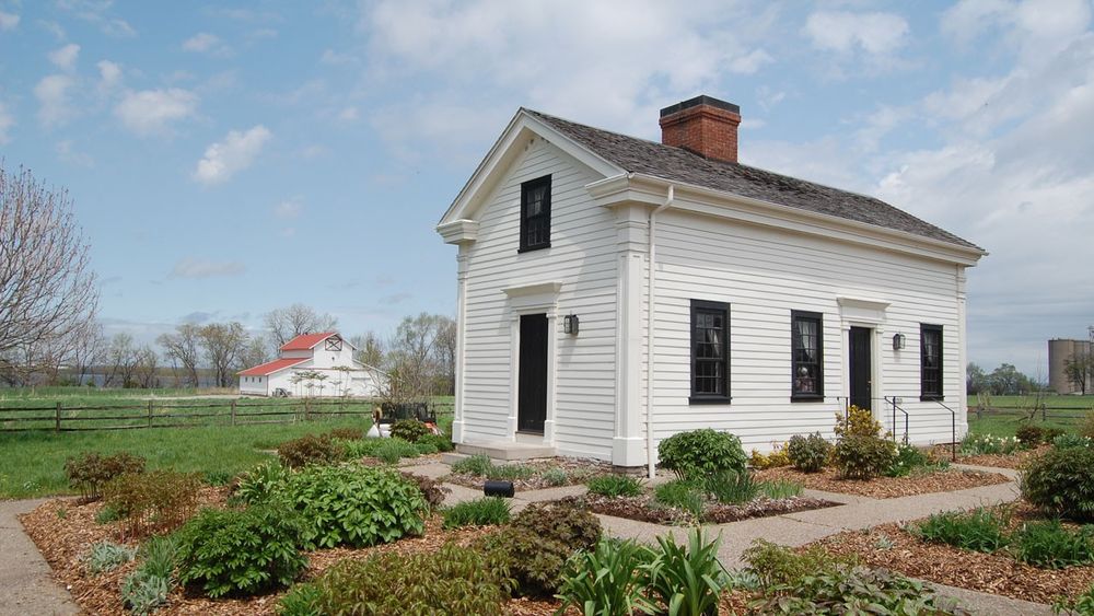 One-and-a-half-story, central chimney, white clapboard home with a well-landscaped surroundings.