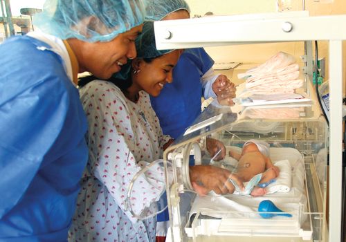 Two doctors in blue aprons looking at a baby as the mother reaches inside the baby incubator in a neonatal intensive care unit in the Dominican Republic.