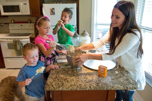 A woman and her three children make cookies in their kitchen, taking spoonfuls of dough out of a large metal bowl.