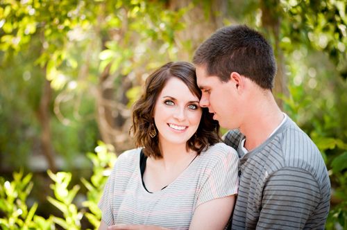 An engagement picture of a young man embracing a young woman while standing outside near overhanging trees.