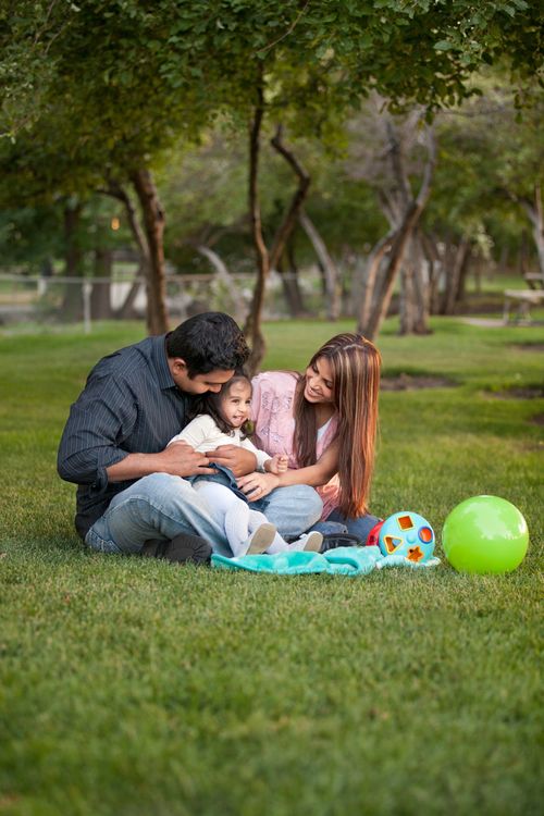 A mother, father, and daughter sit in the park with toys and a blanket.