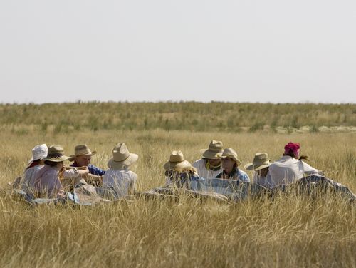 Nine men and women wearing hats all sit together in a grassy field and rest during trek.