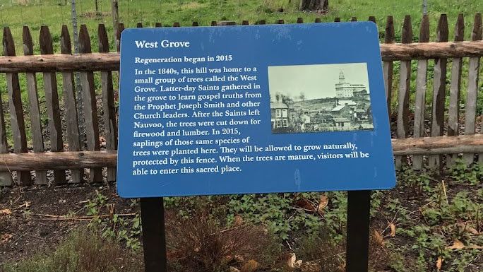 A blue panel that gives history about the West Grove stands in front of a fence with a white temple on the hill in the background.  