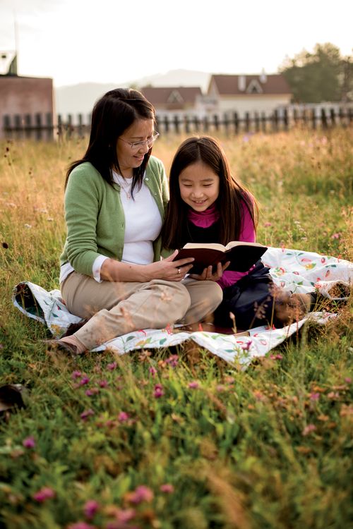 A mother sits next to her daughter on a blanket in a field and holds a set of scriptures open while her daughter reads from them.