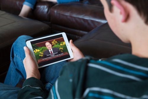 A young man watching general conference on his tablet while sitting on a couch.