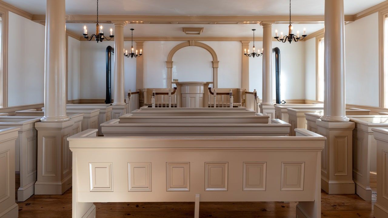 View of large room filled with wooden pews painted white facing a circular rostrum, also of painted wood.