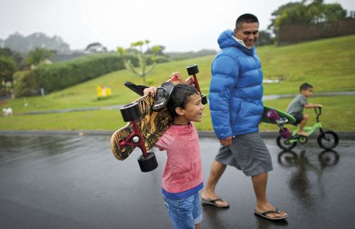 A father and son carrying skateboards as they walk down a wet road.