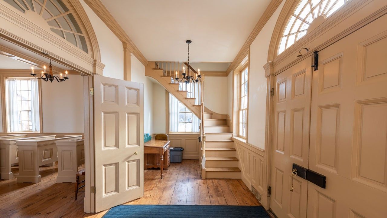 A formal entryway with an L-shaped staircase, closed double doors to the right, and open double doors leading to a large room with pews on the left.