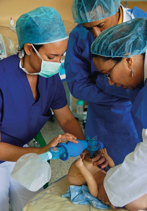 Three doctors in blue shirts and hairnets checking the breathing of a CPR practice baby in the Dominican Republic.