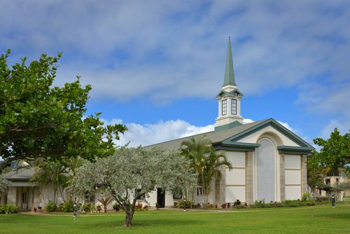 A green lawn and trees in front of a tan chapel with a steeple rising into the blue sky with clouds at BYU–Hawaii.