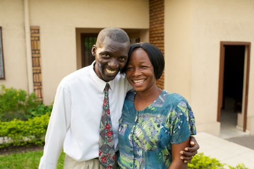 A husband and wife standing and hugging each other outside a building surrounded by green shrubs in Africa.