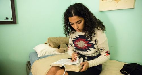A teenage girls sits on her loft bed in her room. She has her feet hanging over the side of the bed. She appears to be reading her scriptures. This is in Maine.