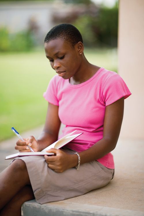 A young woman in Ghana, wearing a pink shirt and a khaki skirt, sits on a step outside and writes with a pen in a journal.
