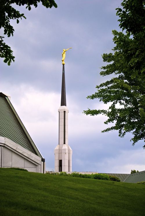 The spire and angel Moroni on the grounds of the Frankfurt Germany Temple, seen over a green hill among large green trees.