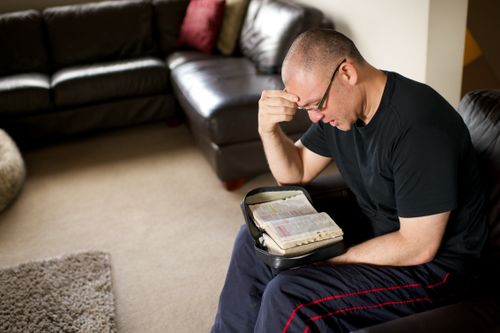 A man sits in an armchair with his scriptures in his lap and reads from them.