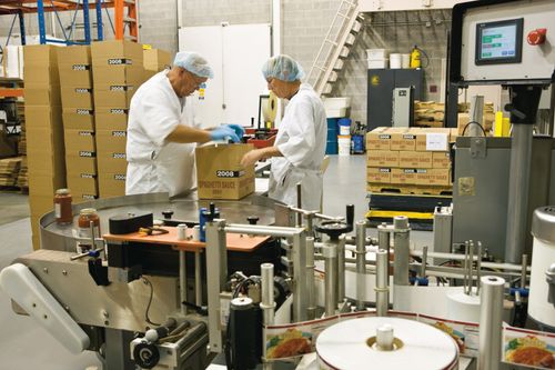 Two men in white aprons, blue gloves, and hairnets closing a box on a food packing line inside of Welfare Square.