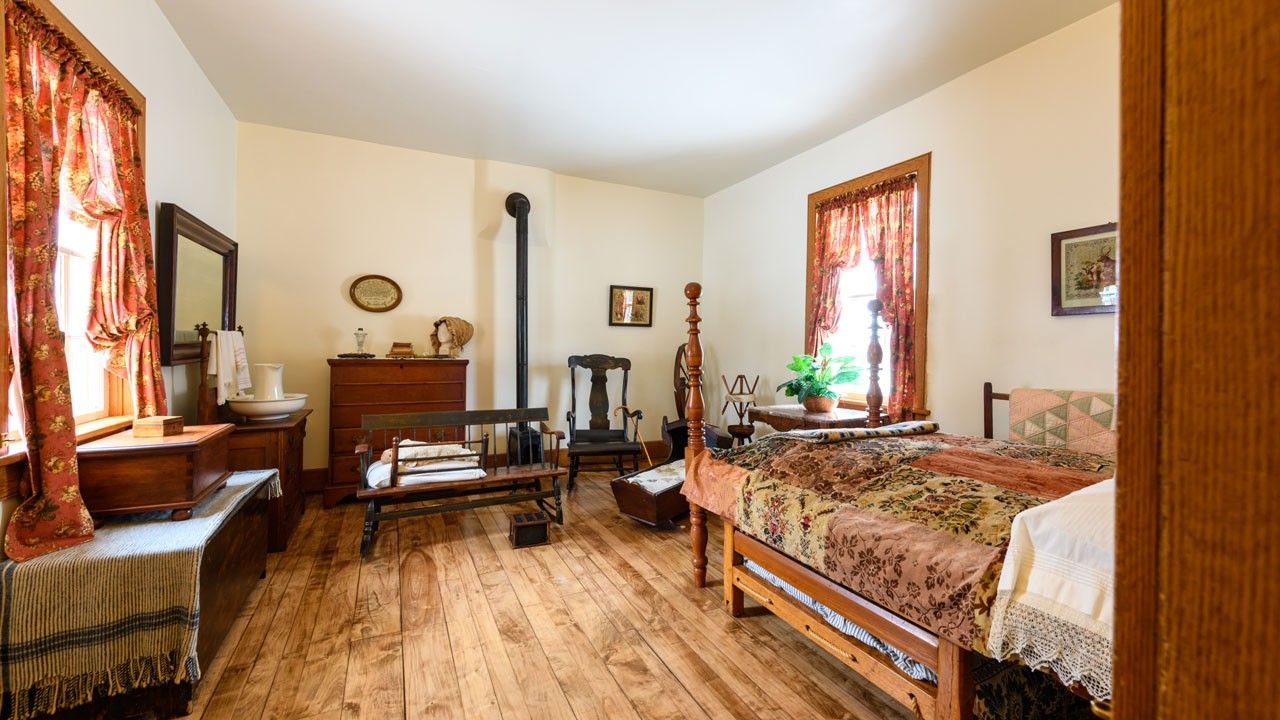 A bedroom interior with wood floor, two windows with red floral curtains, an iron stove, a bed, and other furnishings. 