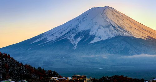 Mt. Fuji, in Japan, rises up majestically in the background on a late autumn evening. The last glimpses of sunlight can be seen on the west facade of the snow capped slopes. Lake Kawaguchi is in the foreground with the town of Fujikawaguchiko nestled in between the lake and mountain. Light snow has blanked the town and the nearby trees.