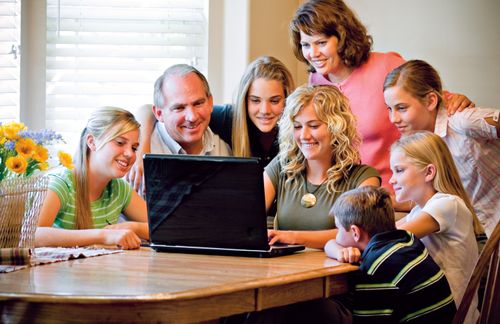 A young woman sits at a dining room table and works on a laptop while her mother and father and five siblings sit around and watch what she is doing.