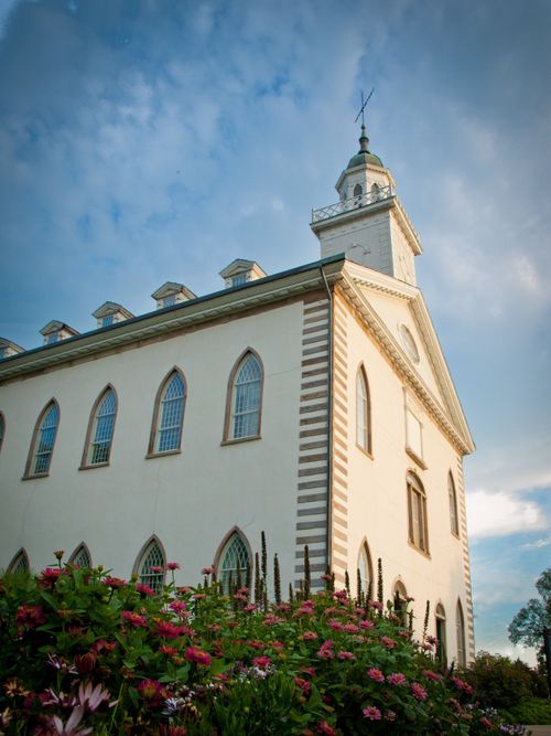 A side view of the front part of the Kirtland Temple, with a green and pink flowering bush in the foreground and light clouds in the distance.