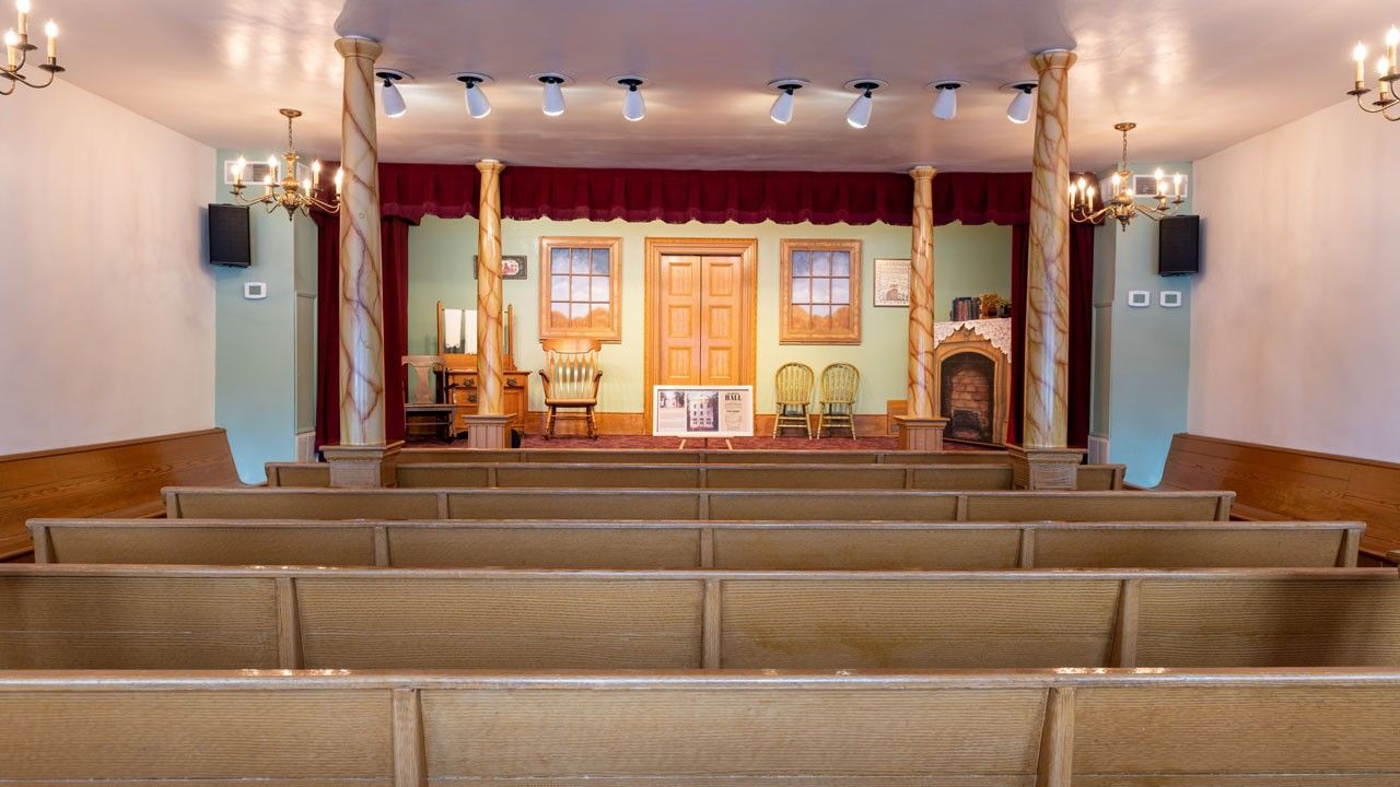 Interior view of building’s main floor. Wooden pews face a stage set for a play. 