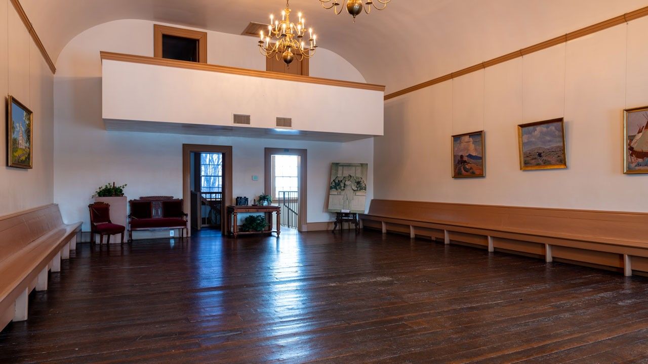 Interior view of the building’s third floor. There are dark wooden floors, a wooden pew on either side of the room, pictures on the walls, and a balcony overhead. 