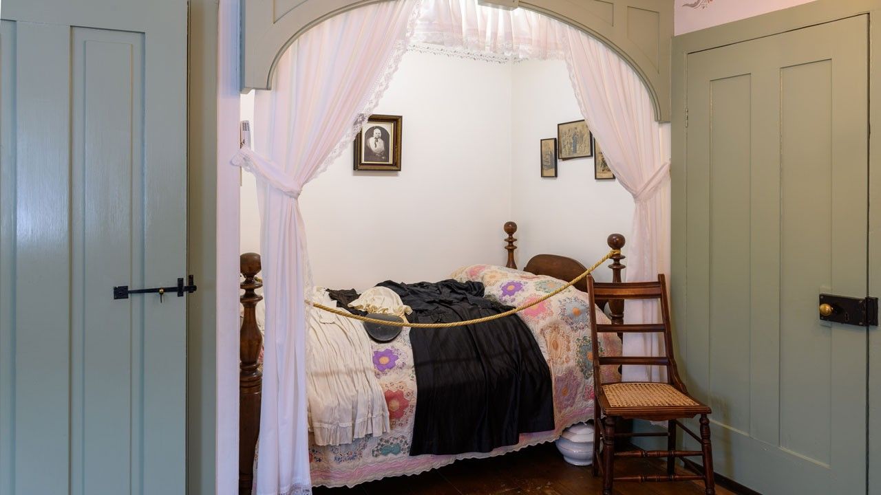 Bed alcove framed by white curtains on either side. A wooden four-poster bed with a floral quilt is tucked into the space.