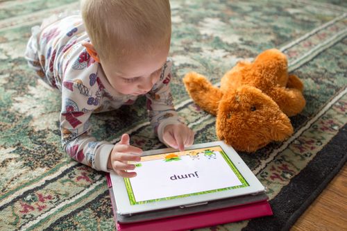 A toddler with short blonde hair and pajamas lies down on a rug beside a teddy bear and plays a word game on a tablet.