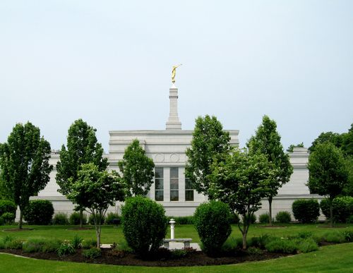 Leafy green trees on the grounds of the Palmyra New York Temple, partially obscuring a view of the temple with the angel Moroni on the spire.