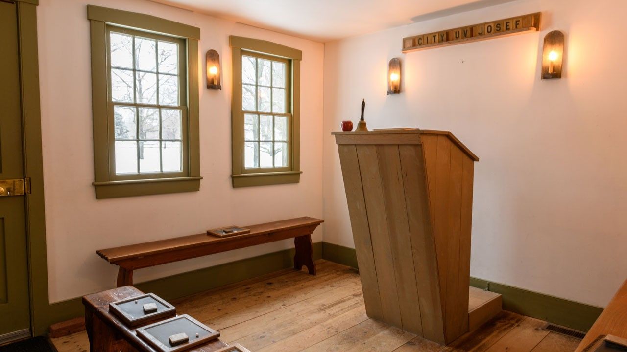 Replica schoolroom with a teacher’s podium and benches. Small blackboards with chalk and erasers are sitting on the benches.