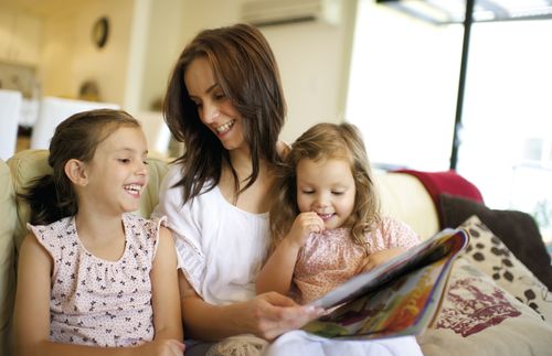 A mother and her young daughters sitting together on a sofa.  They are looking at a magazine.