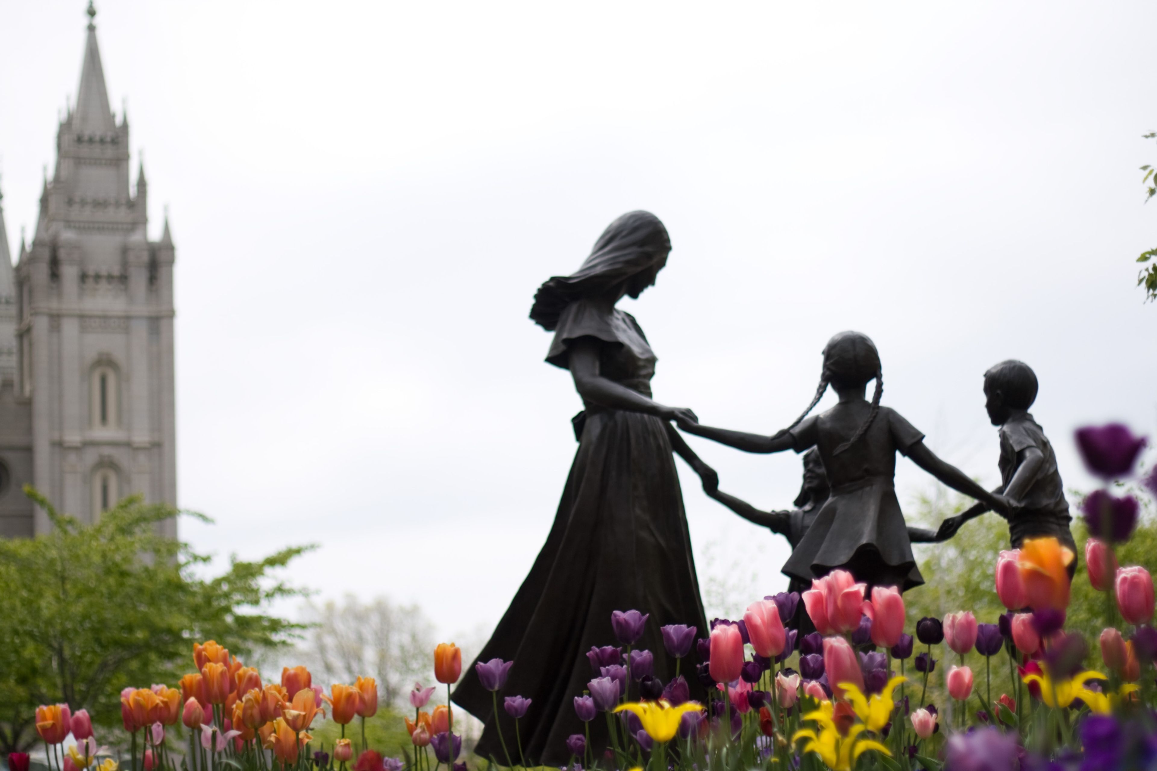 A sculpture of a family on Temple Square has tulips blooming around it, with the Salt Lake Temple in the background.