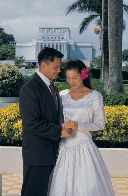 A man in a dark suit and a woman in a white wedding gown standing together in front of the Laie Hawaii Temple on their wedding day.
